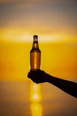 Gün batımında ellerinde bir şişe yaz dönümü temalı bira tutan kadınlar. Bira şişesi güneşten gelen altın ışık, bokeh baloncukları oluşturuyor. Sığ alan derinliği