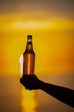 Gün batımında ellerinde bir şişe yaz dönümü temalı bira tutan kadınlar. Bira şişesi güneşten gelen altın ışık, bokeh baloncukları oluşturuyor. Sığ alan derinliği