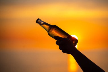 Gün batımında ellerinde bir şişe yaz dönümü temalı bira tutan kadınlar. Bira şişesi güneşten gelen altın ışık, bokeh baloncukları oluşturuyor. Sığ alan derinliği