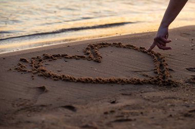 Kadının elini kumsalda parmaklarıyla kalbi çizerken yakından bak. Günbatımında tropik deniz kıyısında romantik aşk sembolü
