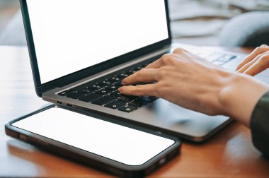 Ahşap bir masanın üzerine akıllı telefon yerleştirilmiş modern bir dizüstü bilgisayarda daktilo eden ellerin yakın çekimi. Her iki ekran da boş, fotokopi alanı sunuyor. Görüntü, uzaktan çalışma, dijital iletişim, üretkenlik, teknoloji ve modern çalışma alanı yaşam tarzını temsil etmektedir..