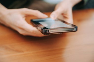 Ahşap bir masanın üzerinde el ele tutuşup akıllı telefon kullanmak. Yumuşak doğal ışık ve sığ alan derinliği ile modern mobil iletişim, çevrimiçi tarama, mesajlaşma ve dijital yaşam tarzı kavramı.