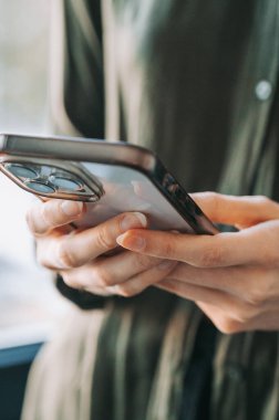 Modern akıllı telefonu kapalı mekanda tutan ve kullanan bir kadının yakın çekimi. Doğal ışık, sığ alan derinliği ve çağdaş tasarım dijital iletişim, mobil tarama ve günlük teknoloji kullanımı.