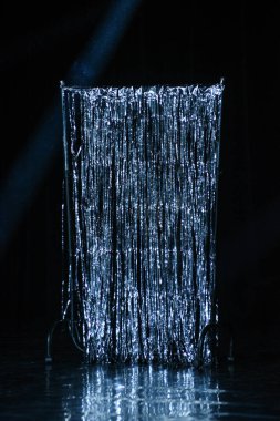 Karanlık bir sahnede odaklanmış bir spot ışığıyla aydınlatılan parıldayan gümüş renkli süs perdesi. Dramatik, yüksek kontrastlı sahne prodüksiyon, parti arkaplanları, tiyatro gösterileri ve eğlence kavramları için ideal.