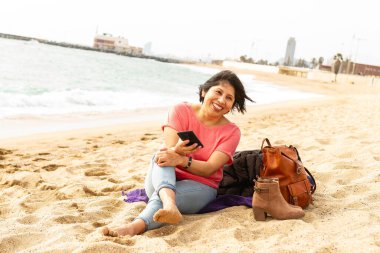 Plajda, havlunun üzerinde çıplak ayaklı bir kız cep telefonuyla video görüşmesi yapıyor.