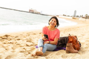 Plajda, havlunun üzerinde çıplak ayaklı bir kız cep telefonuyla video görüşmesi yapıyor.