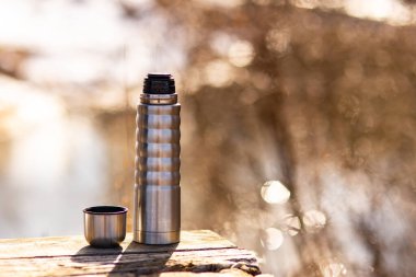 Sıcak çayla termos. Bahar doğası. Nehrin arka planında termos var. İlkbaharda yürüyüş yaparken ısınmaya devam et..