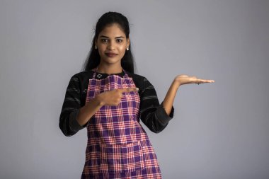 Hintli kadın aşçı portresi ya da önlüklü aşçı, sunar, gösterir, gösterir, işaret eder, tamam işareti ile, gri arka planda baş parmaklar.