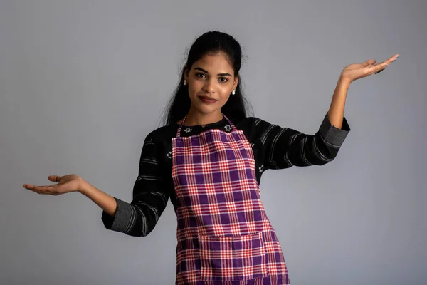 Portrait of Indian woman chef or cook in an apron, presenting, pointing, with ok sign, thumbs up on the grey background.