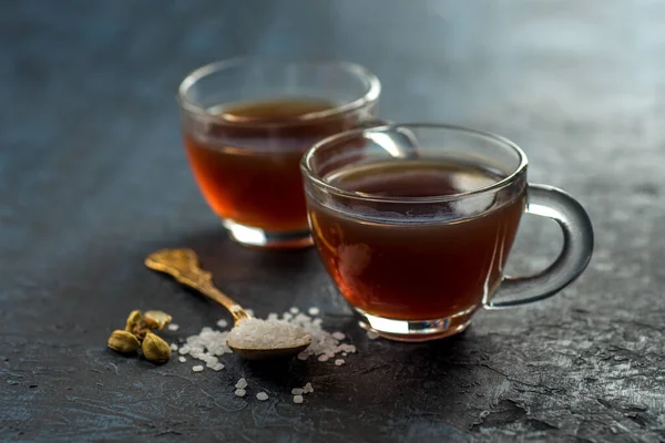 A cup of tea and cardamom with sugar in a spoon on a textured background.