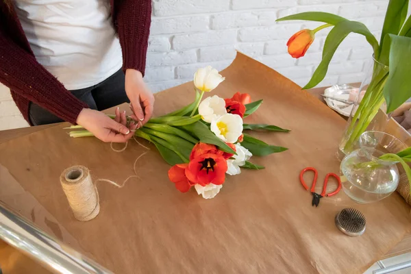 顧客のためのチューリップの創造的な花束の作成に取り組んでいる女性花屋 最上階だ 花屋で新鮮な白と赤のチューリップ 花屋さんの机にハサミと花瓶とセーター — ストック写真