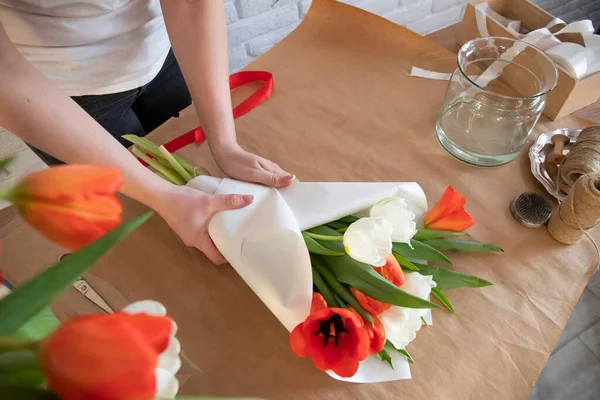 女性は白い紙に赤と白のチューリップの花束を梱包しています 花屋の職場だ 愛する人への贈り物を準備する 春の花のコンセプト — ストック写真