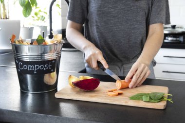 Gri tişörtlü bir kadın modern, parlak bir mutfakta tahta bir tahtada sebze kesiyor. Kova dolusu gübre. Masada artık sebzelerle gübrelemek için metal bir kova. Sıfır kayıp. Geri dönüşüm.