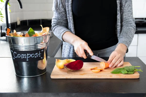 A woman in a black T-shirt cuts vegetables on a wooden board in a modern bright kitchen. Bucket with compost. Metal bucket for composting with residue vegetables on a table. Zero waste. Recycling.