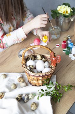 Ahşap bir masada paskalya kompozisyonu. Bıldırcın ve tavuk yumurtası sepette. Kadın arka planda Paskalya yumurtalarını boyuyor..