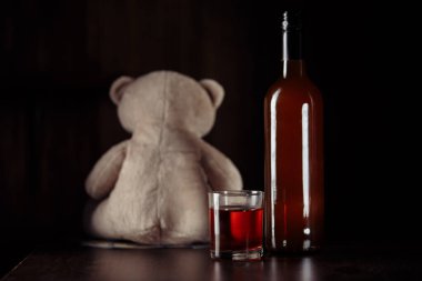 Alkol ve aile kavramı. Oyuncak ayı, çocukların güvenlik sembolü ve karanlık bir odada camla dolu bir şişe.
