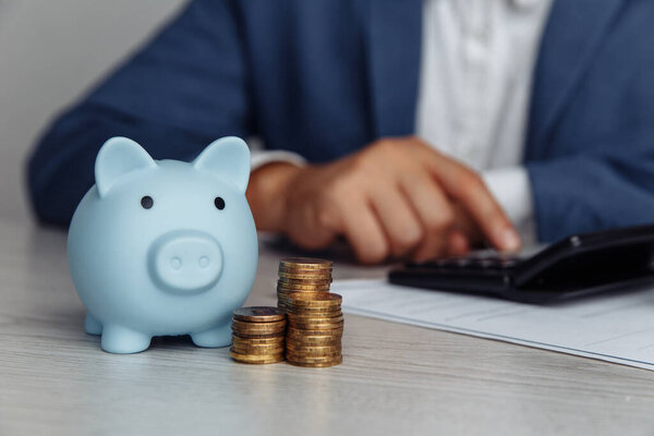 Man is counting profit on a calculator, blue piggy bank with stack of coins on wooden desk in office. Economy and management financial concept