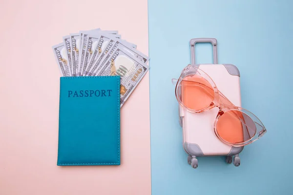 Valise de voyage avec des lunettes de soleil et passeport avec de l'argent sur fond bleu rose. Concept de tourisme — Photo