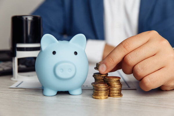 Blue piggy bank, stamp and mans fingers with coins on wooden desk in office. Save money and management financial concept