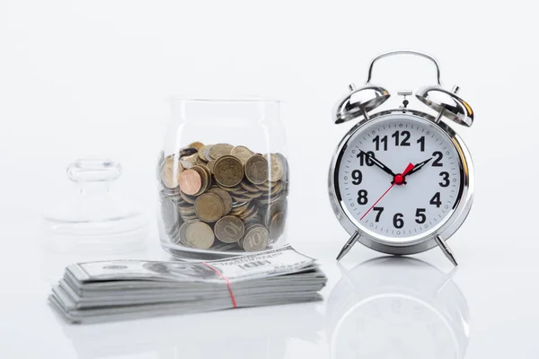 Banco con monedas, billetes de dólar y reloj despertador en una mesa de cristal. El tiempo es concepto de dinero — Foto de Stock