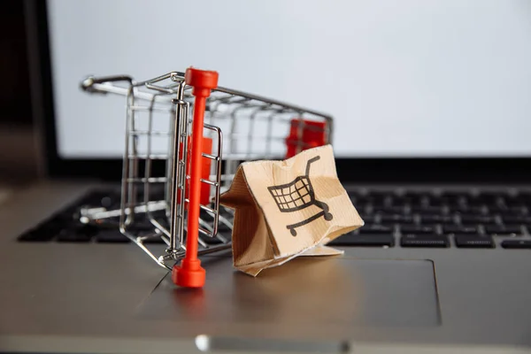 Damaged paper box and shopping cart on a keyboard of laptop. Cheap parcel delivery, poor shipment quality