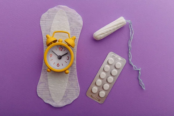 女性の月経周期の概念。薄紫色の背景に衛生パッド、タンポン、目覚まし時計 — ストック写真