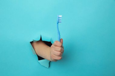Woman hand showing a tooth brush out of a hole torn in blue paper wall. Health care concept clipart