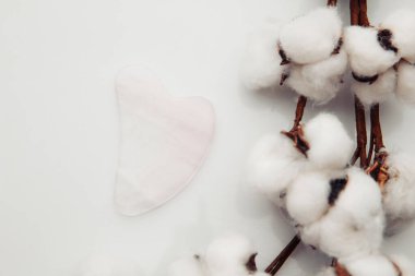 Pembe Gua Sha masaj aleti beyaz bir masada. Evde yüz bakımı, yaşlanmayı önleme ve kaldırma terapisi.