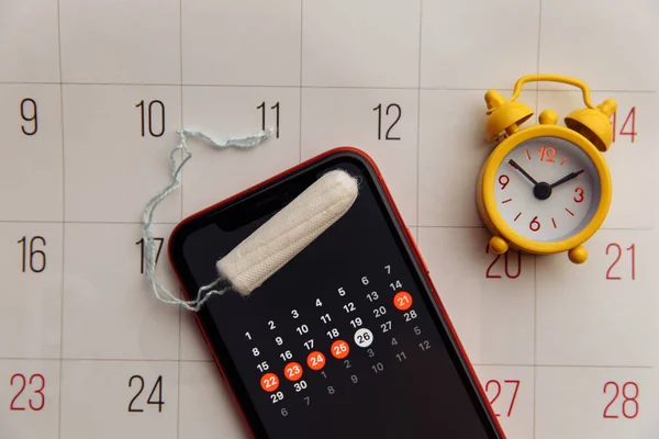 Menstruation app in smartphone with cotton tampon and alarm clock on a calendar close-up. Woman critical days and hygiene protection concept