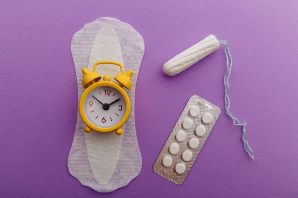 Menstruatiepad, katoenen tampon, pillen en gele wekker op lila achtergrond. Menstruatiecyclusconcept voor vrouwen — Stockfoto