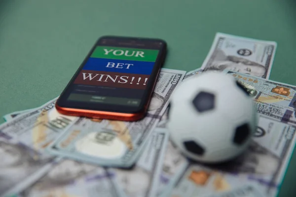 Concepto de negocio de fútbol. Pelota y smartphone con aplicación de apuesta en billetes de dólar y fondo verde. concepto de dinero de juego de fútbol — Foto de Stock