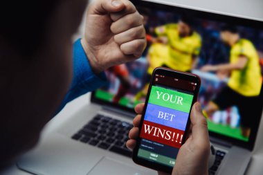 Futbol seyreden bir adam dizüstü bilgisayarında yayın yapıyor ve bahisçilerin web sitesinde bahis oynayarak zaferi kutluyor. Mesajlı akıllı telefona odaklan