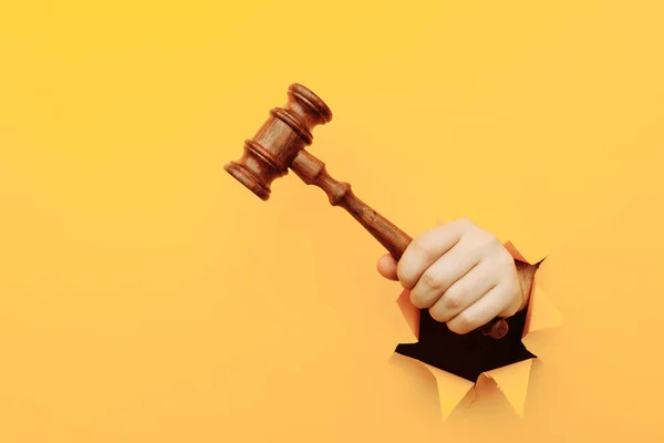 Hand holding a judges gavel through torn yellow paper wall. Law and courts, triumph of justice — Stock Photo, Image