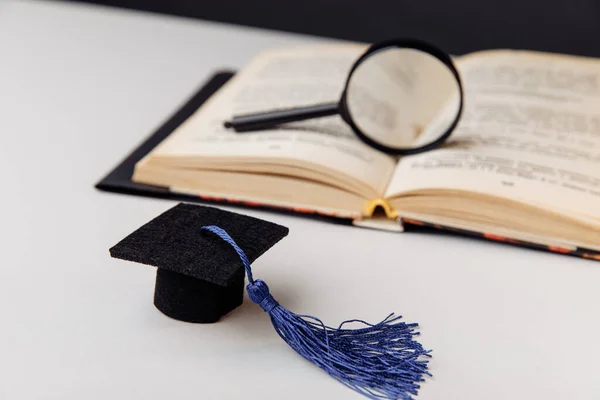 Lupa en libro abierto y gorra de graduación en mesa blanca — Foto de Stock