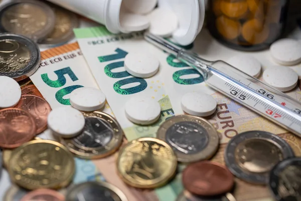 Pills and medicine with clinical thermometer on money. Euro in banknotes and coins. One hundred Euro bill in focus