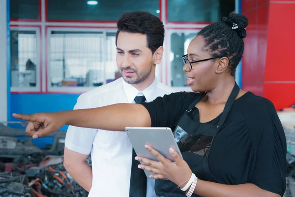 Diversity of two people, black African worker woman explain detail of product on a tablet to caucasian business manager in factory-warehouse