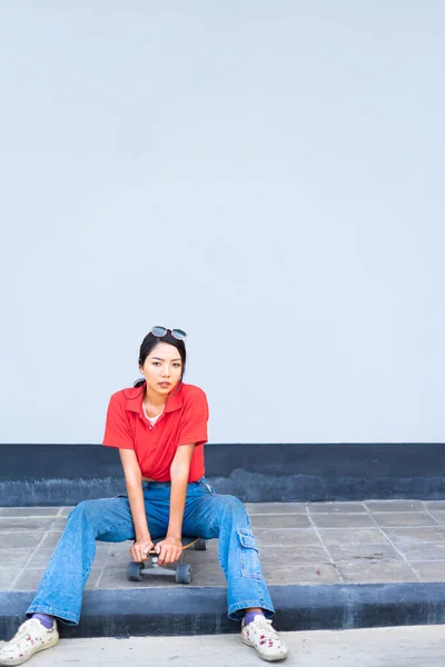 Asian cute girl or woman sit on skateboard on left side, looking straight, and act smart at cool wall with copy space in summer holiday.