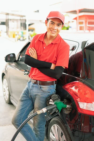 Asian gas station worker man leaning against black car with happiness while green fuel nozzle filling high energy power fuel into black auto car tank in petrol station, commercial service for benzine, diesel, gasohol, gasoline.