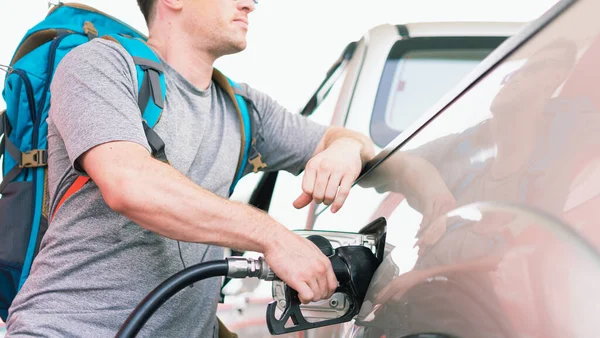 White caucasian car owner with blue backpack holding black fuel nozzle and filling high energy power fuel in auto car tank in petrol station, commercial service for benzine, diesel, gasohol, gasoline.