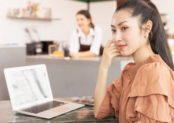 Beautiful Asian woman customer stop using laptop and looks at left side while waiting for coffee in coffee shop. Barista work at coffee bar and food service business