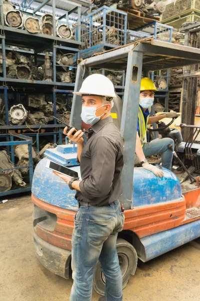 Men work together, wear safety facemask, use walkie-talkie. Caucasian engineer man wear safety facemask and use walkie-talkie while Asian man driving forklift in factory-warehouse