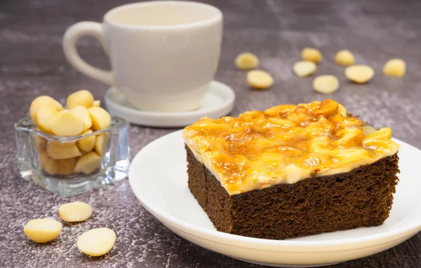 Toffee chocolate cake slice on plate with macadamia seeds and coffee cup on stone background for dessert break. Toffee cake made from bake, dark chocolate, macadamia, nut, caramel, and coffee