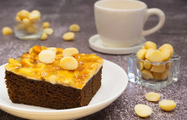 Toffee chocolate cake slice on plate with macadamia seeds in small bowl and coffee cup on stone background for dessert break. Toffee cake made from bake, dark chocolate, macadamia, nut, caramel
