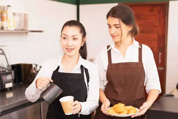 Focusing on half-body angle of Asian barista women stop filling milk into takeaway hot coffee cup while talking with customer in cafe coffee shop. Barista work at coffee bar and food service business