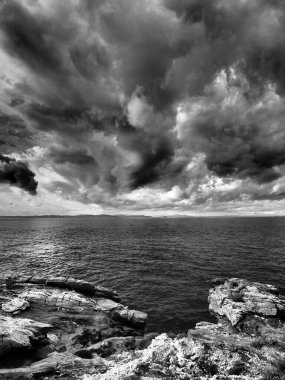 Cyclades 'teki Kea Adası' nın kayalık kıyı şeridinde çekilmiş yüksek kontrastlı siyah-beyaz bir deniz manzarası. Ağır, katmanlı fırtına bulutları açık Ege ufkunun üzerinde gökyüzünü süpürürken, desenli dalgalar ve engebeli kayalıklar ön tarafı kaplıyor.