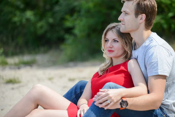 Молодая пара сидит на песке улыбаясь и обнимаясь — стоковое фото