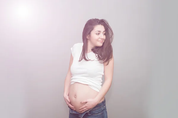 Беременная девушка в расстегнутых джинсах и белой футболке держится за голый живот. Студия на сером фоне — стоковое фото