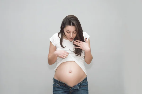 Ragazza incinta in jeans sbottonati e t-shirt bianca aggrappata alla pancia nuda. Lo studio con sfondo grigio — Foto Stock