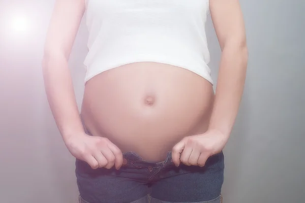 Pregnant girl in unbuttoned jeans and white t-shirt holding on to naked belly. The studio with gray background — Stock Photo, Image