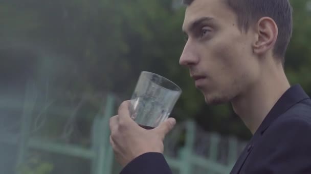 Een jonge man in een jasje staat en drinkt whisky uit een glas — Stockvideo